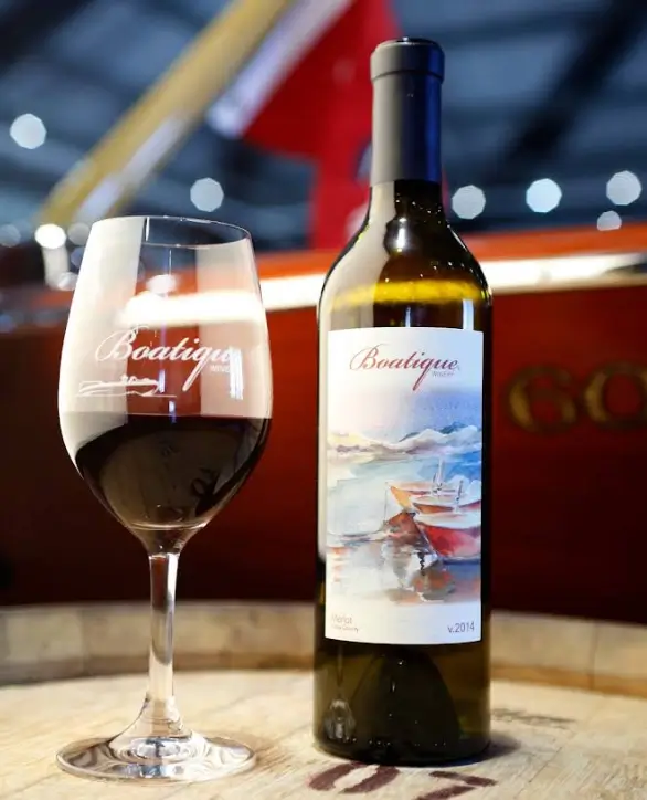 A bottle of Boatique wine and a filled wine glass placed on a barrel with a boat in the background