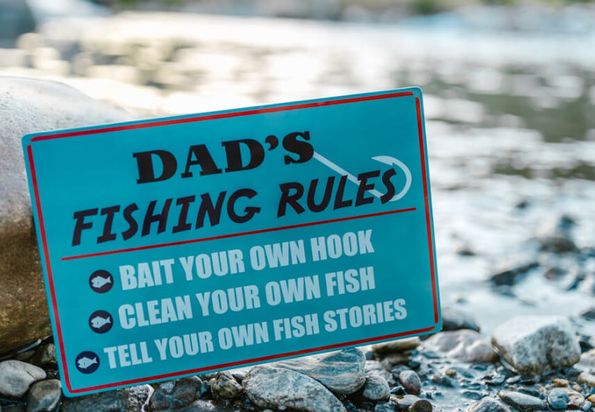 Blue sign by the river that says “Dad’s Fishing Rules”