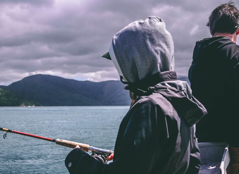 Person in a hooded jacket fishing on a lake with mountains in the background