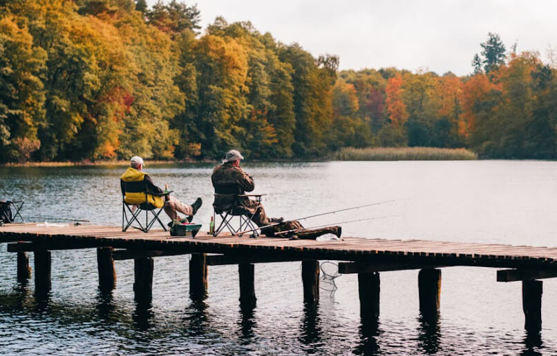 Two people fishing on a wooden dock in front of autumn trees