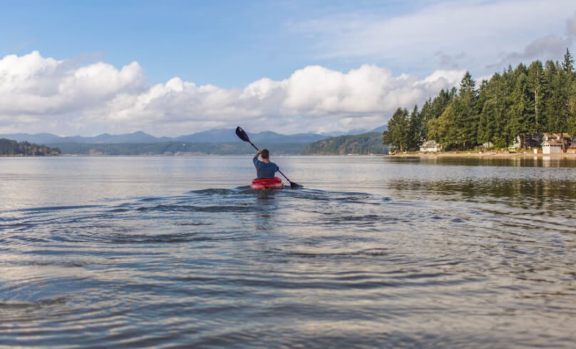 Person kayaking alone on a calm lake with forest and mountains in the distance