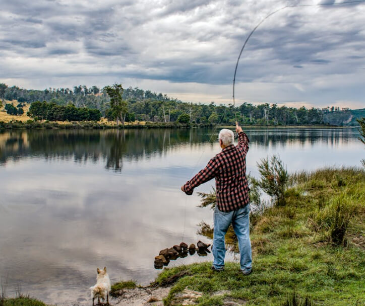 Man fly fishing by a lake with a small dog nearby